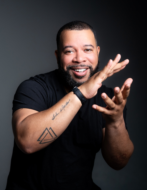 Johnson—a medium-skinned Black man with short dark hair and a close-cropped beard, wearing a black t-shirt—smiles at the camera, his hands caught mid-clap.